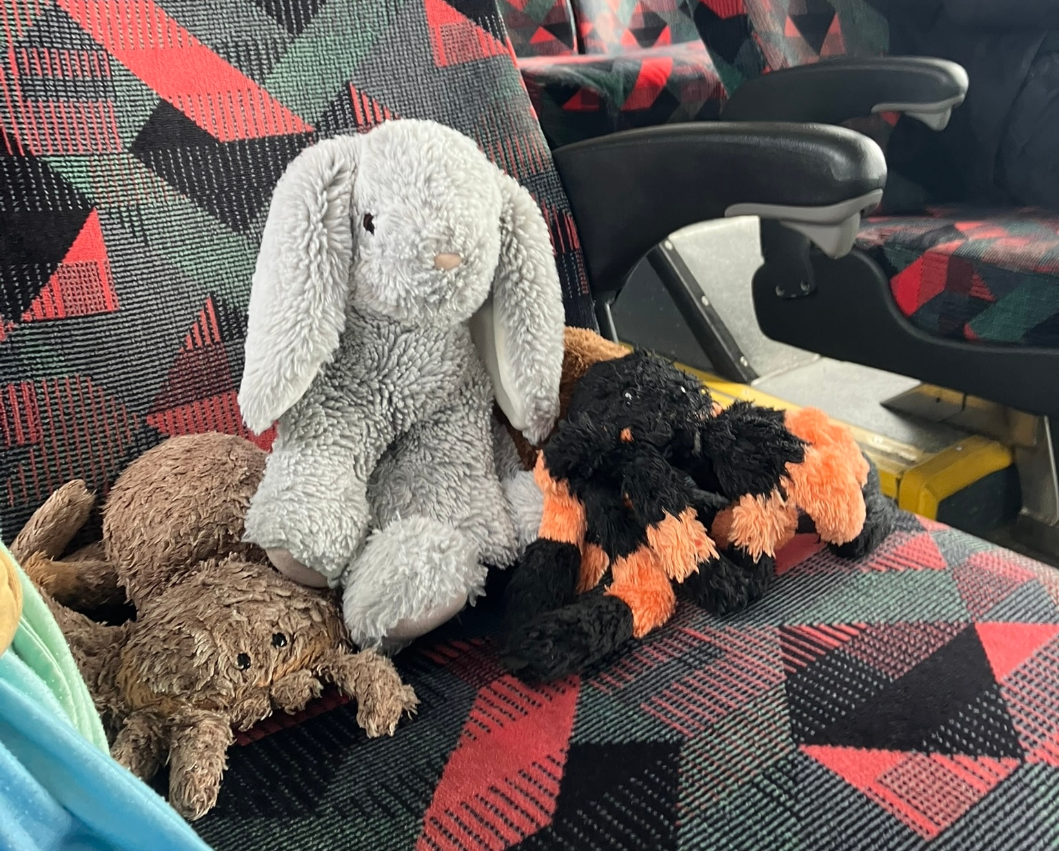 two plush spiders, one brown and one black-and-orange, and a grey stuffed bunny with floppy ears, sit on a bus seat together.